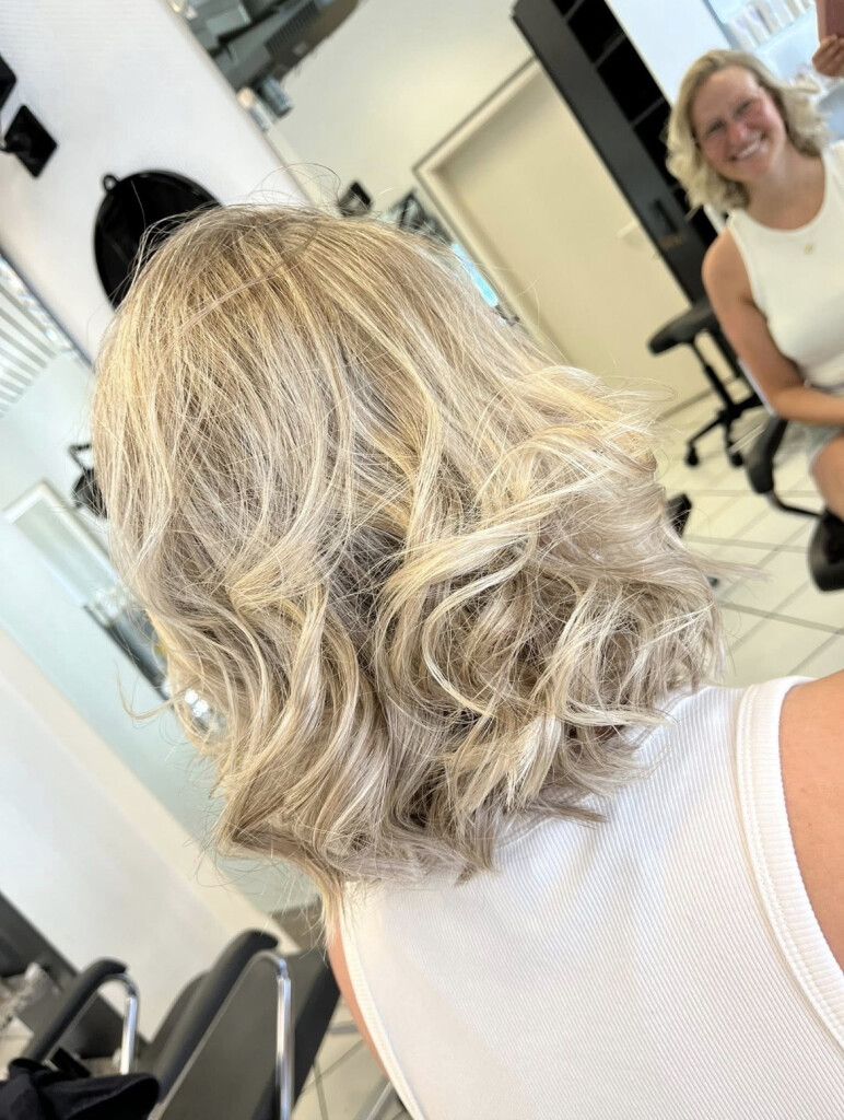Blonde Balayage-Locken von hinten, im Hintergrund lächelt eine Stylistin
